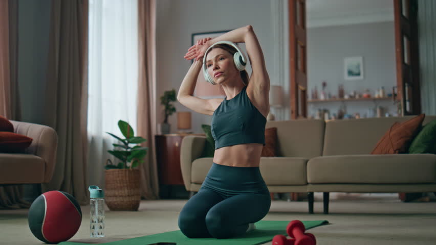 Fit woman warming up body wearing headphones at sport mat home. Focused athletic sportswoman stretching muscles sitting on knees at living room. Sporty girl in sportswear workout in wireless earphones