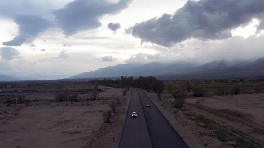 Aerial view scenic shot, cars crossing desert area on straight freeway in rural central Asia, Kyrgyzstan. Asphalt highway and mountains with clouds on horizon, background.