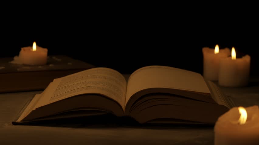 Close shot of retro hardcover book on dark background in warm light. Wind turns dusty pages of vintage book surrounded by candles.