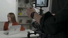 The videographer adjusts the camera, zooming in on the presenter sitting at a table in front of a laptop. The female presenter listens to the instructions before filming and nods her head