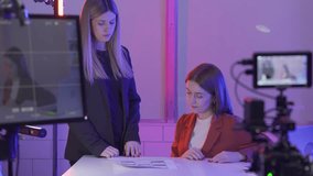 A female presenter discusses a storyboard with a female producer before filming begins in close up. Women in a studio in pink neon light in front of a professional camera.