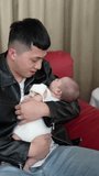 Vertical slow motion video of a Taiwanese mother and father in their 20s giving milk to a 1-month-old girl's baby on a red sofa indoors