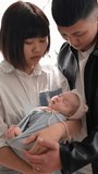Vertical slow-motion video of a Taiwanese mother and father in their 20s standing and toasing a baby girl who is 1 month old in front of the curtain