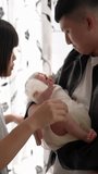 Vertical slow-motion video of a Taiwanese mother and father in their 20s standing and toasing a baby girl who is 1 month old in front of the curtain