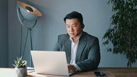 Competent korean businessman with wireless headset conducting video call on modern laptop while sitting at personal desktop in cabinet with blue walls. Male having online conversation with colleagues.