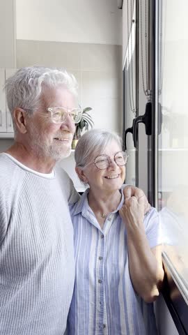 Happy bonding loving middle aged senior retired couple standing near window, looking in distance, recollecting good memories or planning common future, enjoying peaceful moment together at home.
