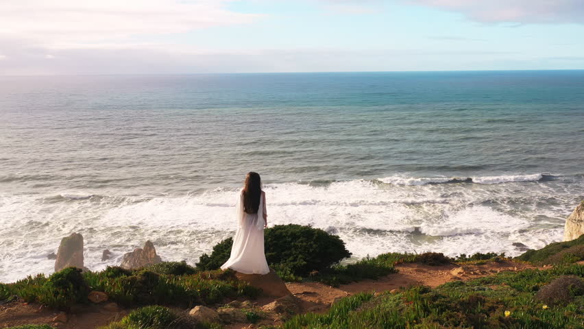Drone rotating around woman in white dress standing on green hill by seascape. Carefree female is enjoying scenic view of vast ocean during vacation. 