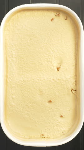 Vertical video - Closeup overhead shot of a hand holding a freshly opened tub of salted caramel ice cream with toffee pieces, while a spoon tool scoops and lifts away a spoonful.