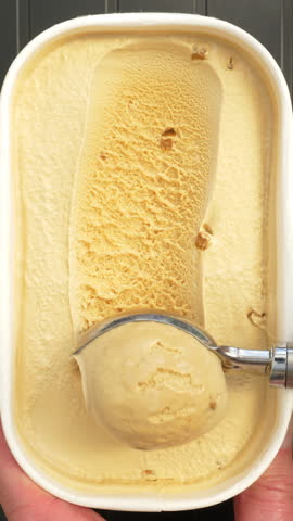 Vertical video - Closeup overhead shot of a hand holding a freshly opened tub of salted caramel ice cream with toffee pieces, while a spoon tool scoops and lifts away a spoonful.