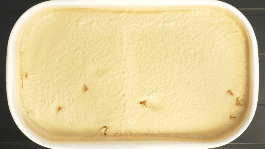 Closeup overhead shot of a hand holding a freshly opened tub of salted caramel ice cream with toffee pieces, while a spoon tool scoops and lifts away a spoonful.