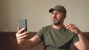 A man with a beard dances. Holding a black smartphone in his hand. Takes a selfie. Records videos for social networks. Green baseball cap and T-shirt. Military. In room. Sitting on a brown sofa agains