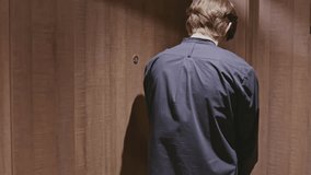 Rear view of a young man closing the door of a hotel room. Stock clip. Man in black clothes in dark interior of hotel room.