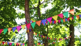 Colorful flags hanging on branches of trees as festive decoration for outdoor party. Bright summer sunshine among green leaves in background. Real time full hd video footage. Christmas, birthday fun