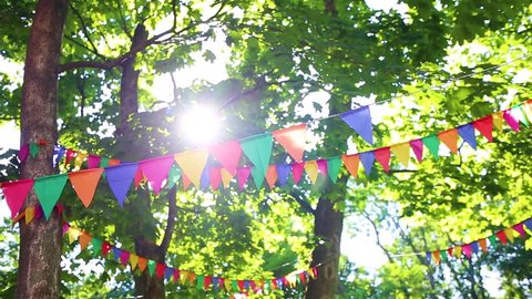 Colorful flags hanging on branches of trees as festive decoration for outdoor party. Bright summer sunshine among green leaves in background. Real time full hd video footage. Christmas, birthday fun