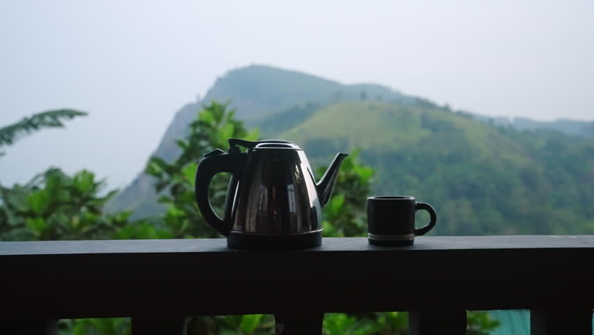 Dolly shot of tea kettle, cup on railing of luxury terrace with mountain view, Ella Rock, Sri Lanka in background. Tea experience, scenic nature at high-end vacation home, serene plantation landscape.