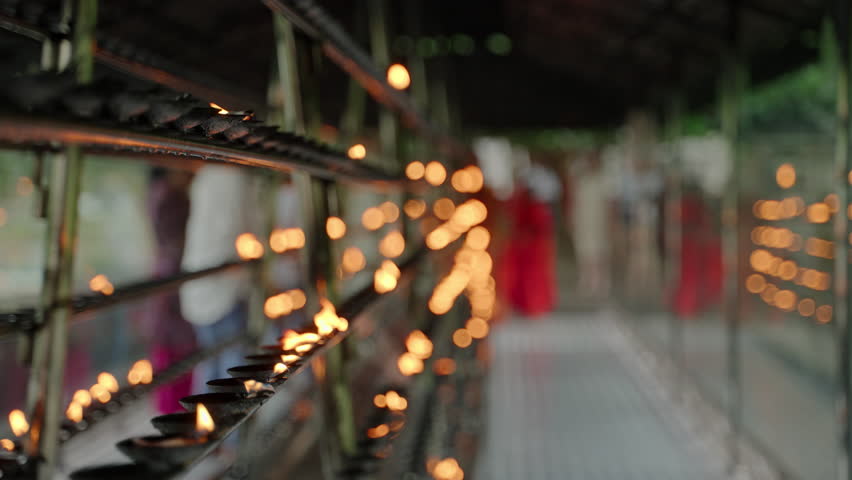 Buddhist devotees light oil lamps, offer prayers in temple. Rows of flickering flames line racks with blurred figures moving, serene religious practice in sacred place, creating peaceful atmosphere. Royalty-Free Stock Footage #3495511065