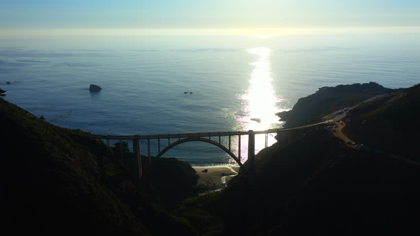 4k cinematic aerial scenic sunset. Panoramic aerial view over arch Bixby Creek Bridge against the backdrop of the blue Pacific Ocean. The sun sinks into the waves on the horizon. Nature of Big Sur.  Royalty-Free Stock Footage #3495595667