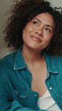 Vertical video, Smiling young woman with curly hair wearing denim shirt looking through window while relaxing on sofa in cozy living room