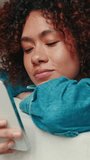 Vertical video, Close-up of young woman with curly hair wearing denim shirt using mobile phone while lying on sofa in cozy living room
