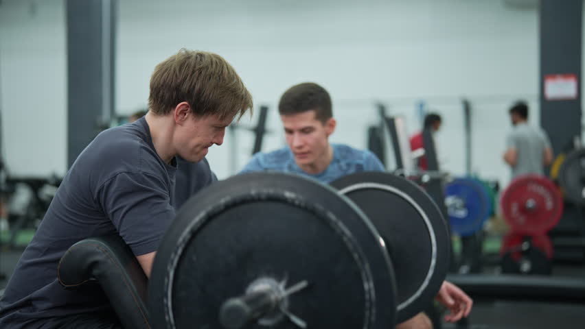 Personal trainer helping beginner sportsman for correct doing Scott Curls with barbell, lifting up EZ-bar sitting on preacher curl bench. Man exercising support under supervision of personal trainer