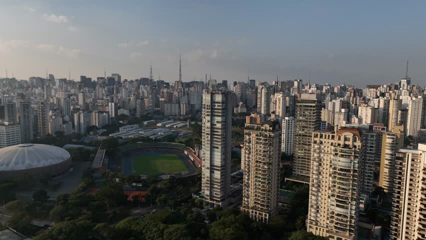 Sao Paulo, Sao Paulo, Brazil, June 7 2023: Aerial view drifting slowly left above Ibirapuera Park sports complex, showing an athletics stadium with running track and nearby domed arena surrounded by trees and residential towers. The Sao Paulo skyline stretches across the background, under clear daylight with radio masts visible, while streets and gardens frame the venues in the foreground, emphasizing the dense urban landscape around the park. Long shadows fall across the field and stands below