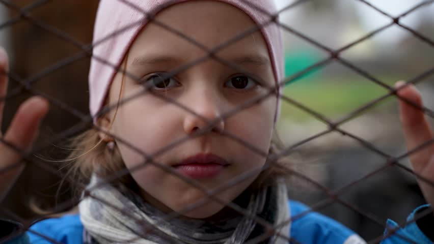Close-up shot of a little girl standing behind wire fence. Portrait of a sad girl behind a fence