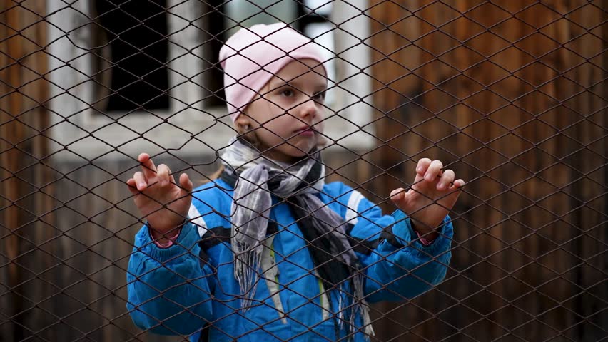 Close-up shot of a little girl standing behind wire fence, selective focus. Portrait of a sad girl behind a fence