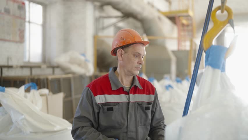 Industrial worker in safety gear operating a crane hook of cathead, crane in factory. Moving heavy bags in the workshop. 