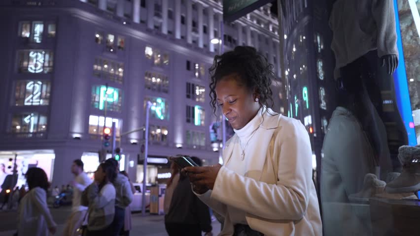Young latina woman waits sitting for her date in night illuminated winter city center. Smiling afro female types on mobile phone in dating app. Generation z black people and social relationships. Royalty-Free Stock Footage #3496468765
