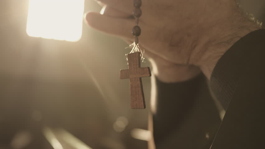 Christian rosary with cross hanging from male hands and swinging close up. Monk holding prayer beads in clasped arms while praying in cathedral closeup. Christianity concept