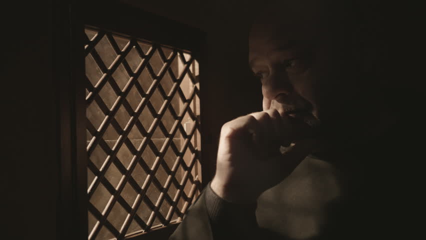Mid adult monk sitting in confessional booth near window in church close up. Caucasian Christian priest in monastery robe listening to confession in cathedral
