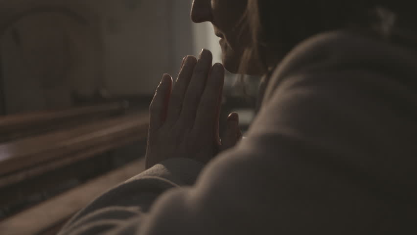 Folded arms of religious woman praying in Christian church close up. Mid adult female sitting with closed eyes during prayer in cathedral side view. Catholicism concept