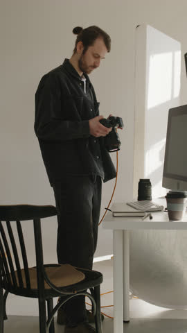Vertical medium shot of young Caucasian male photographer looking at photos on digital camera display while working in studio