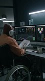 Full vertical of African American male postproduction studio employee with mobility impairment sitting in wheelchair at desk with dual monitor, checking script and editing raw video for movie