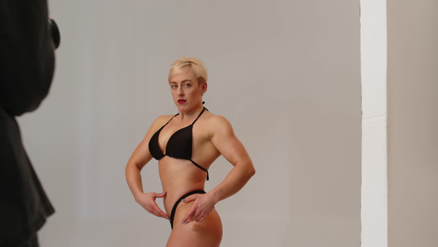 Medium shot of blonde Caucasian fitness bikini model posing for camera on white studio background and photographer taking pictures on digital camera