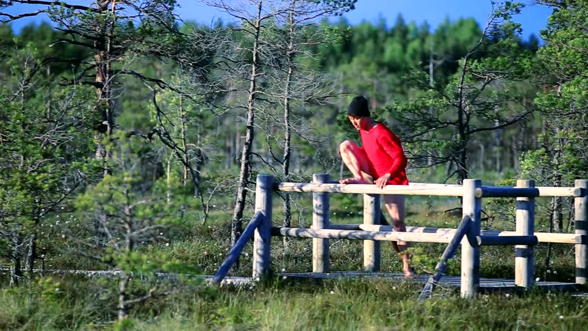 Chinese contemporary dancer in red shirt and black hat is performing on the path among the bog in National Park of Kemeri. Side view.