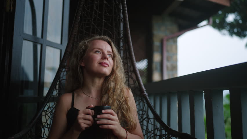 Woman relaxes in swing chair on scenic terrace, sips hot tea amidst mountain views, tea gardens at dawn. Serene, she enjoys warm brew, tranquil atmosphere on cozy patio, overlooking rich landscape.