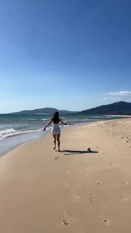 A girl in a white dress is running along the beach. Enjoy the sunny day and the sea. Vertical