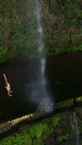 Vertical video. Aerial view of happy woman traveler in white swimsuite near a waterfall in the highlands, raises his hands up, enjoys nature and life.Lifestyle concept. Madeira island, Portugal