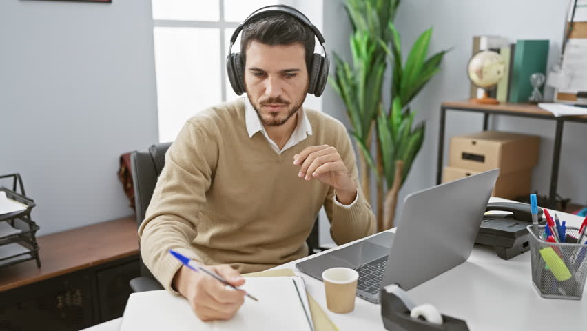 A contemplative young hispanic man with headphones and beard working on a laptop in an office setting takes notes.