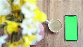 Close-up of a smartphone with green screen near cup with coffee on flowers background. Press browsing chroma key online, typing text, reading social media. Slow motion
