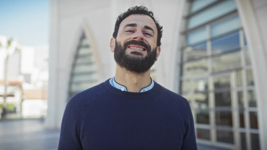 Smiling bearded man in casual clothing standing outdoors with modern architecture in the background