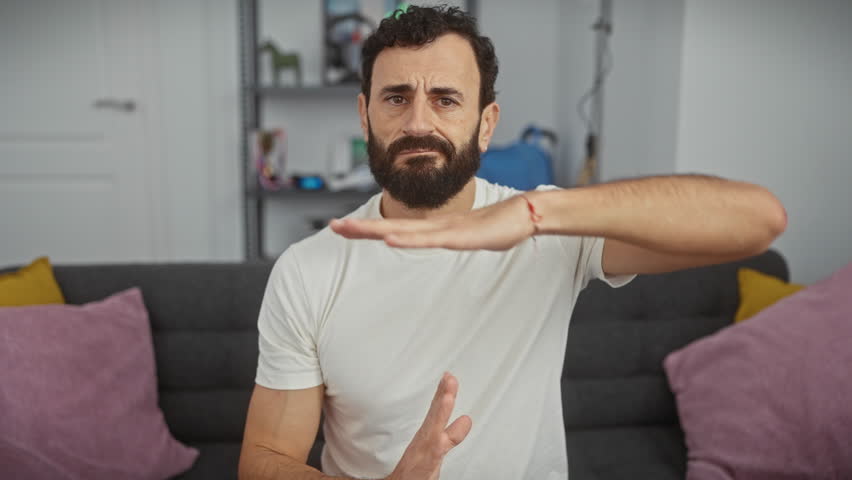 Frustrated middle age man, confidently wearing a white t-shirt, showing a time out gesture with his hands, his face all serious, full of arguments in his indoor home setting.