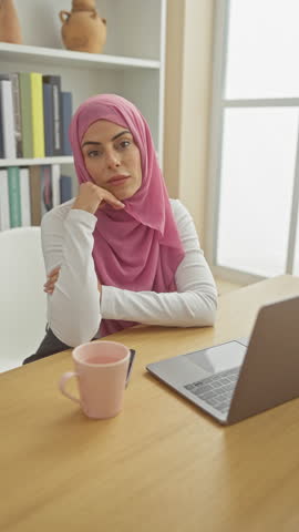 A thoughtful woman with a hijab sits at a desk with a laptop and a pink mug, in a cozy, bookshelf-lined room. Royalty-Free Stock Footage #3497255635