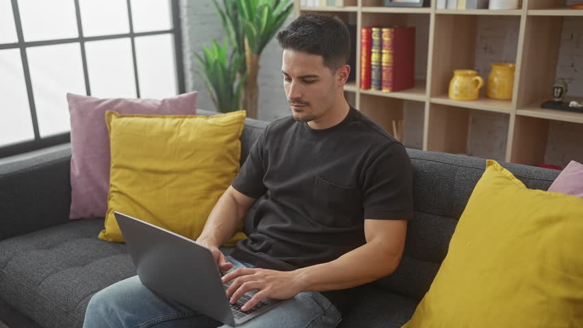 A young hispanic man with a beard working on a laptop in a modern living room with colorful cushions and bookshelves. Royalty-Free Stock Footage #3497266411