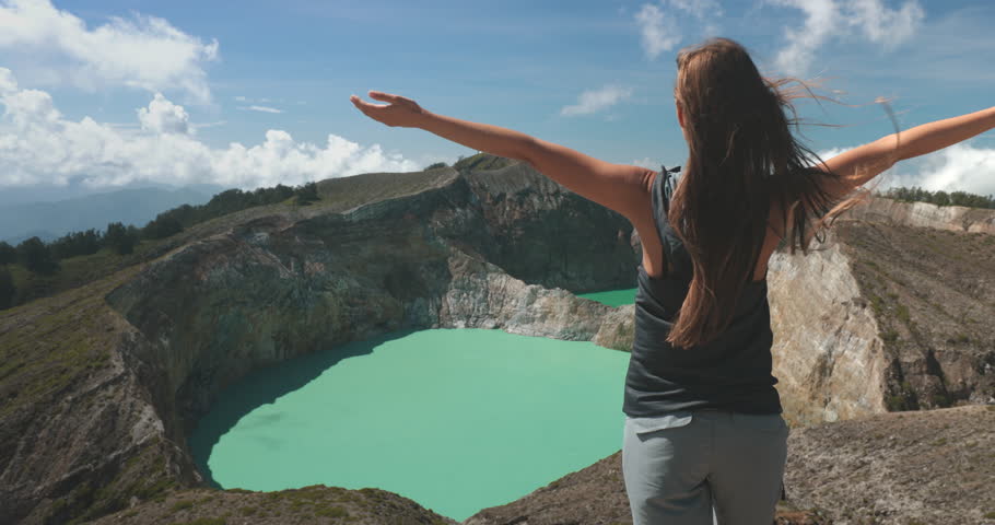 Tourist woman rises hands on volcanic Kelimutu blue crater lakes. Travel destination. Beautiful wild nature landscape. Exotic summer vacation, hiking, relax. Close up back view 4K zoom in