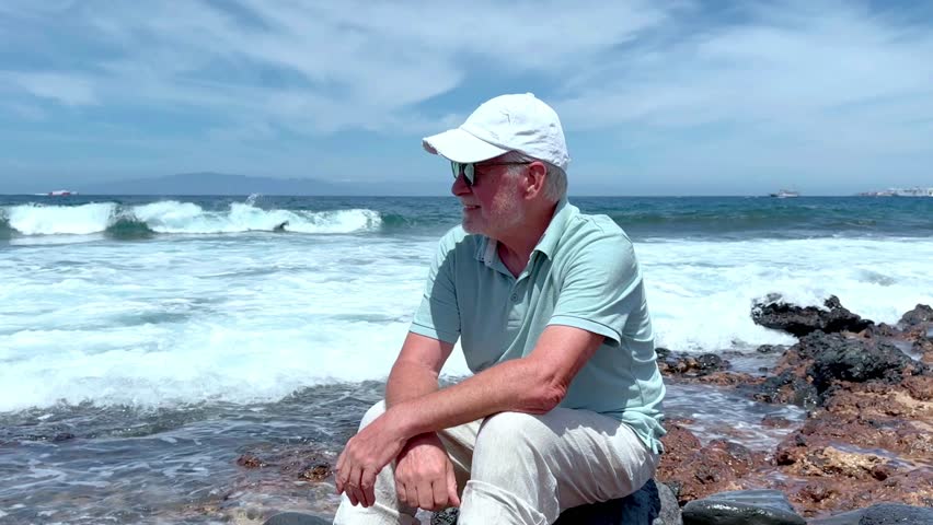 Senior white haired man sitting barefoot at the rocky beach looking the waves crashing on the rocks in a windy day. Elderly man in retirement or vacation enjoying sunny day and freedom