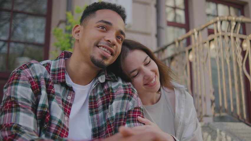 Multi-ethnic couple in love sit on the steps of their home or hotel and enjoying the moment. The guy and the girl are happy together, talking and smiling at each other