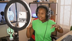 African american woman streaming in a gaming room with led lights, wearing headphones, and using a ring light.
