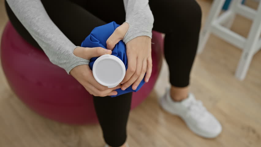 A youthful woman seated on a stability ball in a clinic, focusing on her physical recovery, holding an ice pack.
